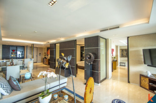 Interior of a modern, spacious apartment with open-plan design, featuring a living area with a plush L-shaped sofa, contemporary art pieces, a large flat-screen TV, and a partition leading to a dining space. The decor includes cultural sculptures, elegant flower arrangements, and a palette of neutral tones with accents of black and gold.